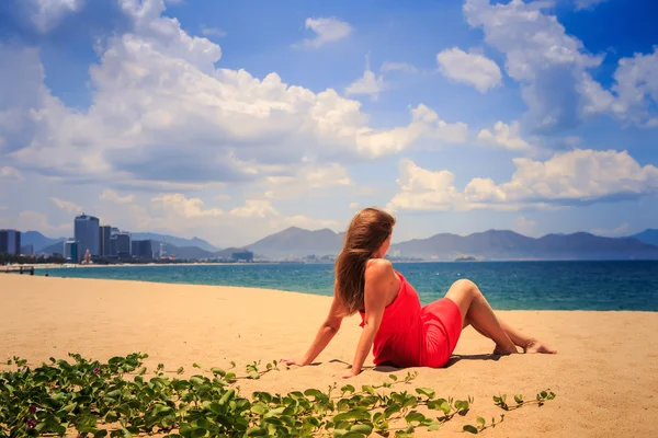 Chica en rojo se sienta en la arena mira al mar en primer plano enredaderas —  Fotos de Stock