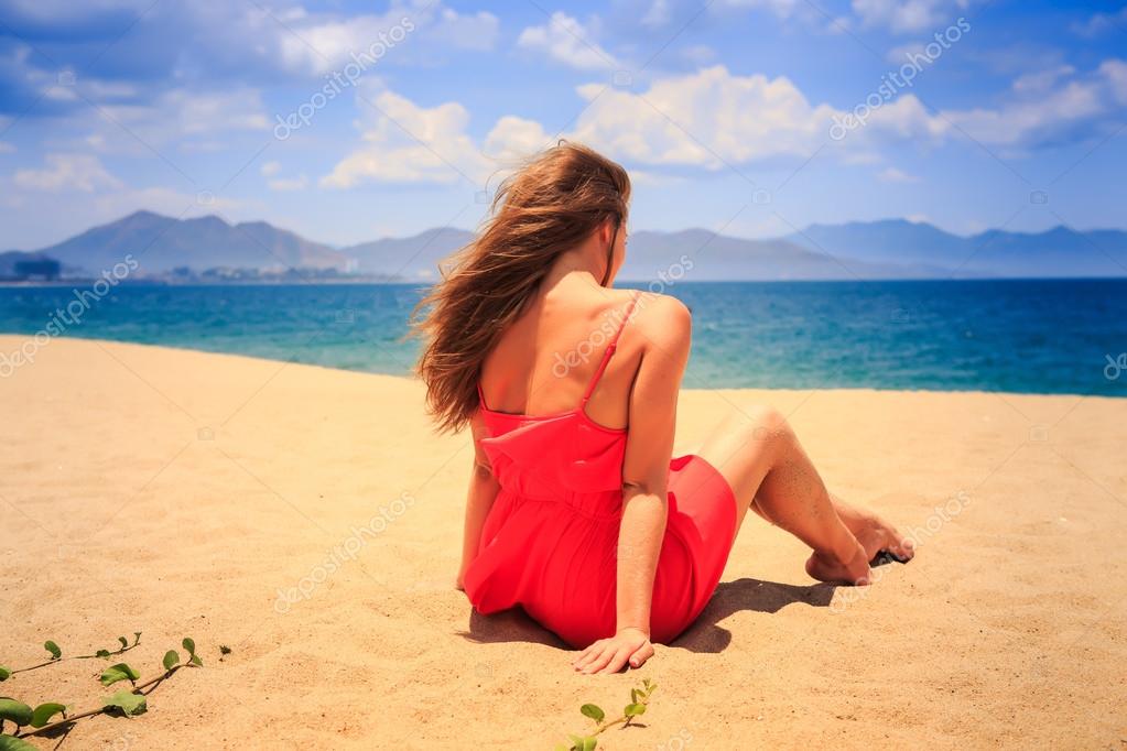 Mädchen in rot mit nackten Rücken sitzt auf Sand sieht auf See