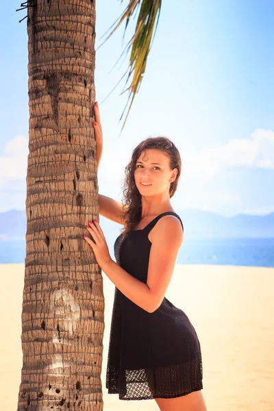 Slim girl in black lace frock barefoot touches palm on beach — Stock Photo, Image