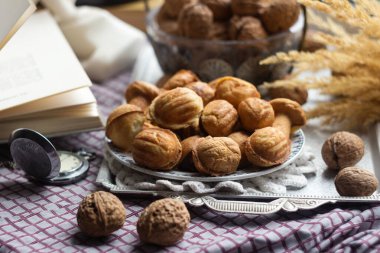 Kareli bir masa örtüsünün üzerindeki metal tepside ceviz şeklinde kurabiyeler, yanında gerçek ceviz kabukları, açık bir kitap ve diğer dekorlar var..
