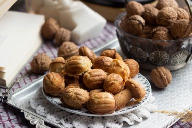 Kareli bir masa örtüsünün üzerindeki metal tepside ceviz şeklinde kurabiyeler, yanında gerçek ceviz kabukları, açık bir kitap ve diğer dekorlar var..