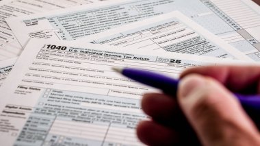 Vergi formlarını bir ofis masasında kalem kullanarak dolduran biri. Formlarda 2025 yılı için 1040 bireysel gelir vergisi iadesi yer alıyor. Evraklar masanın üzerine serildi..
