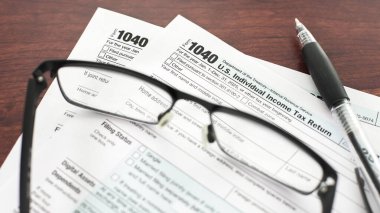 ABD gelir vergisi beyannamesi 1040 ile ilgili belgeler, okuma gözlüğü ve kalemle birlikte ahşap bir masanın üzerinde duruyor..