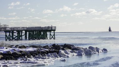 NORWALK, CT, ABD - 30 HAZİRAN 2026: Donmuş bir Long Island Sound açık bir gökyüzünün altında uzanıyor. Ufukta bir deniz feneri dururken, tahta bir iskele suya doğru uzanır. Kar yakındaki kayaları kaplıyor..