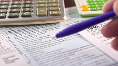 Bir kişi bir masada 1040 vergi formunda çalışır. Sayılar ve hesaplamalara yardımcı olmak için bir hesap makinesi kullanıyorlar. Vergi bilgileri ve talimatları gösteren belgeler dağıtıldı..