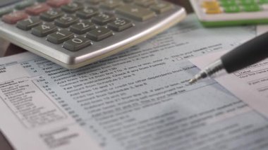 Bir kişi ev ofis ortamında 1040 vergi formu hazırlamak için çalışır. Bir hesap makinesi ve çeşitli kağıtlar masaya serilmiştir, bu da vergi işlemini gösterir..