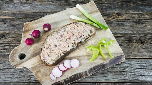 Tütsülenmiş alabalık ile kaplanmış taze dilimlenmiş ekmek ahşap bir kesme tahtasına konur. Yakınlarda, yeşil soğanlar, turplar ve yeşil dolma biber dilimleri renk ve tazelik ekler..