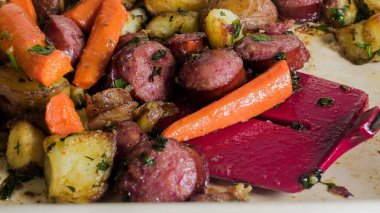 Dilimlenmiş sosis ve kızarmış sebzeler fırın tepsisine konur. Havuç ve patates otlarla karıştırılır. Sahne hazır yiyecekleri gösteriyor, canlı renkleri vurguluyor..