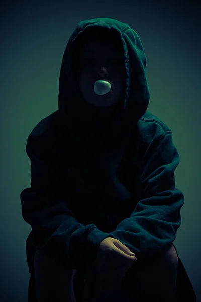 在黑暗的环境中 一个穿着帽衫吹泡糖的孩子的画像 图库照片