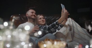 Mutlu genç bir aile, akşamları evdeki deri koltukta otururken akıllı telefondan selfie çekerek eğleniyor. Yakışıklı adam ve gülümseyen kadın cep telefonuyla fotoğraf çekiyor.