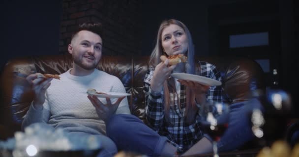 Piękna para siedzi na kanapie i bawi się oglądając wieczorem program telewizyjny lub film w domu. Przystojny mężczyzna atrakcyjna kobieta jedząc pizzę, rozmawiając uśmiechając się podczas dyskusji, co widzą na ekranie — Wideo stockowe