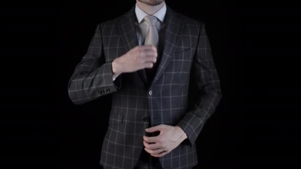 Bonito homem de negócios vestindo terno endireita sua gravata com a mão isolada no fundo preto. Vista sem rosto de jovem caucasiano vestindo blazer xadrez terno de negócios olhando para a câmera — Vídeo de Stock