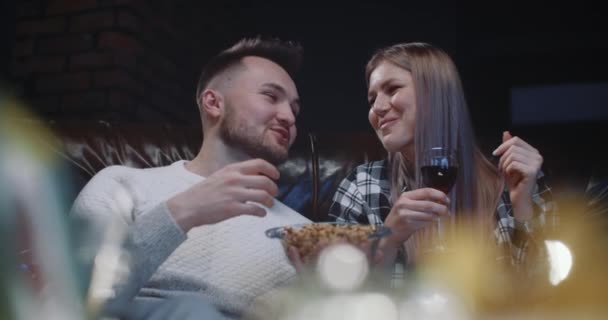 Happy millennial couple sitting on couch having fun watching TV show at home in evening. Handsome man and beautiful cheerful woman eating snacks, drinking wine, talking nicely and smiling each other — Stock Video