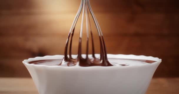 Primer plano mezclando chocolate líquido derretido con batidor de acero en la cocina. Mezcla de chocolate caliente remolino y goteo en un tazón blanco sobre fondo de madera. Cocinar postre de chocolate hecho a mano. Concepto de confitería — Vídeo de stock