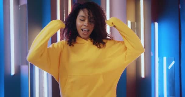 Young cheerful african american girl wearing yellow hoodie dancing and having fun on colourful neon lights background. Happy stylish woman dancing modern energetic dance and smiles emotionally — Stock Video