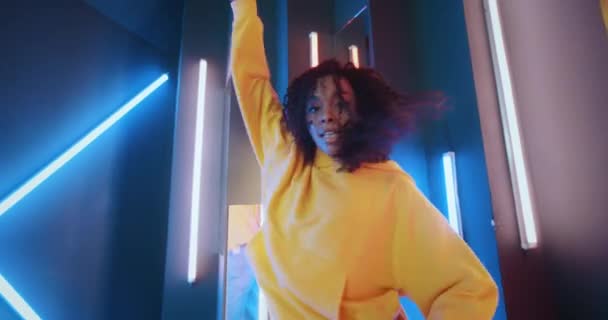 Geniş açı pozitif Afro-Amerikan siyah kadın sarı kapüşonlu gece kulübünde mor ve mavi neon çizgili ışıklarla dans ediyor. Mutlu genç şehir kadını dans eden modern anlamlı dans — Stok video