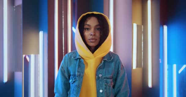 Retrato de mujer negra afroamericana urbana con estilo en sudadera con capucha amarilla y chaqueta jeans de pie en discoteca nocturna mirando a la cámara retroiluminada con luces de rayas de neón púrpura y azul — Vídeo de stock