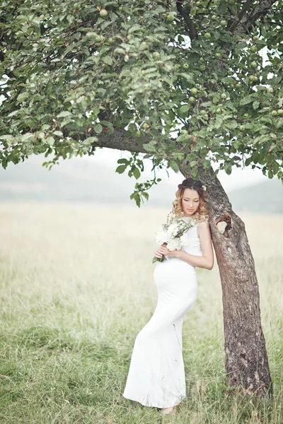 Zwangere vrouw genieten van de zomer park, het dragen van lange witte jurk, holding in handen boeket van bloemen, buitenshuis, nieuw leven-concept. verse stemming — Stockfoto