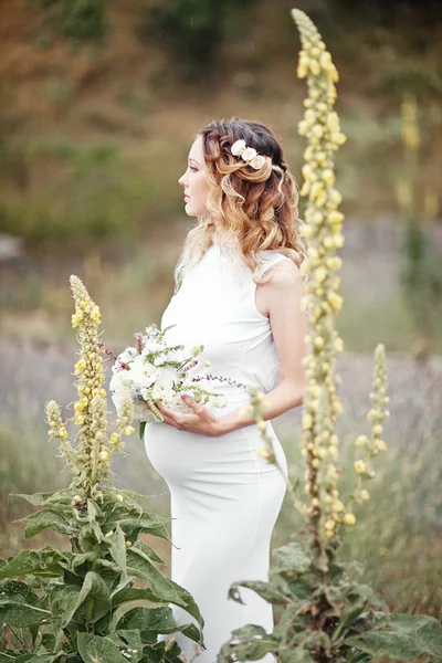 Mulher grávida desfrutando de parque de verão, vestindo vestido branco longo, segurando em mãos buquê de flores, ao ar livre, novo conceito de vida. humor fresco — Fotografia de Stock