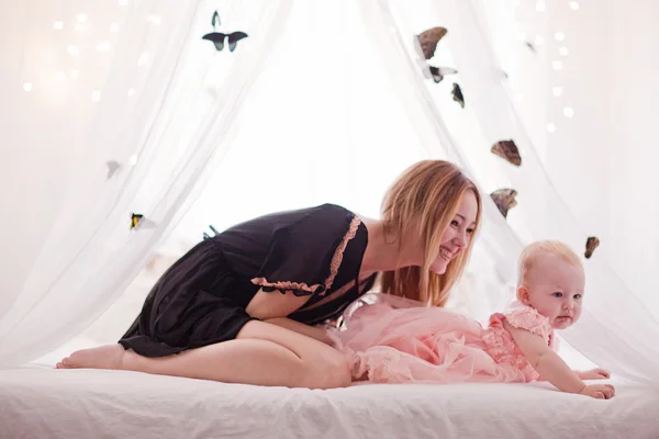 Hermosa joven con hija pequeña sentada en la cama blanca con mariposa . —  Fotos de Stock