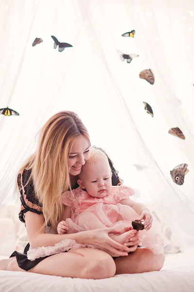 Belle jeune femme avec petite fille assise sur le lit blanc avec papillon . — Photo