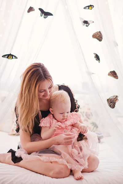 Hermosa joven con hija pequeña sentada en la cama blanca con mariposa . —  Fotos de Stock