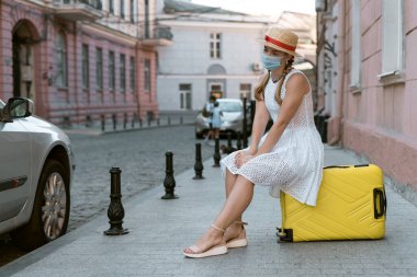 Genç bir kadın büyük sarı bir bavula oturur. Yüzüne tıbbi maske takılmış..