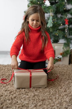 Avrupalı görünümlü kız kırmızı bir kazak giyiyor. Hediye kutusuna fiyonk bağlar