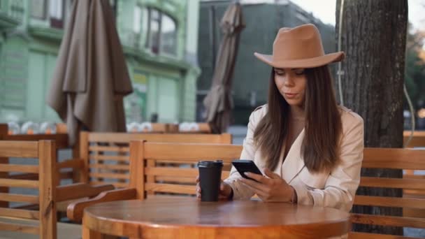 若い美人が通りのカフェに座っている。使い捨てのコーヒーカップを保ちなさい。スマートフォンで番号をダイヤルします。 — ストック動画