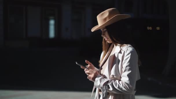 Glad ung kvinna tittar på skärmen på sin smartphone och ler när du går längs gatan i den gamla staden. — Stockvideo