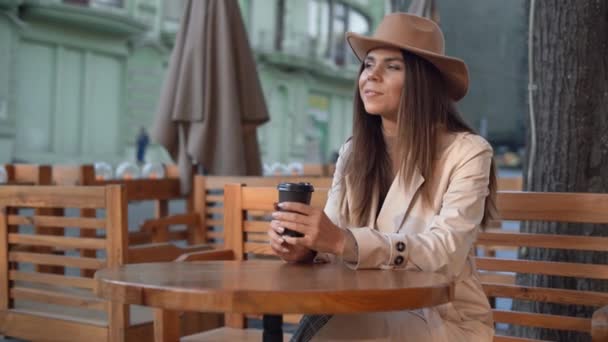 Jonge mooie vrouw met een vilten hoed zit aan een tafel in een café op straat. koffie drinken, wegwerp papieren beker — Stockvideo