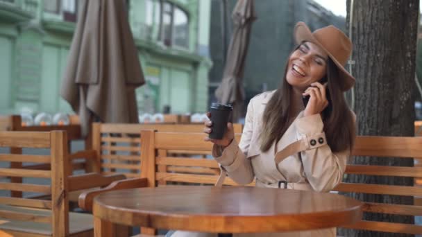 年轻美丽的欧洲姑娘坐在街上一家咖啡馆的桌子旁。快乐的微笑，在电话里交谈 — 图库视频影像