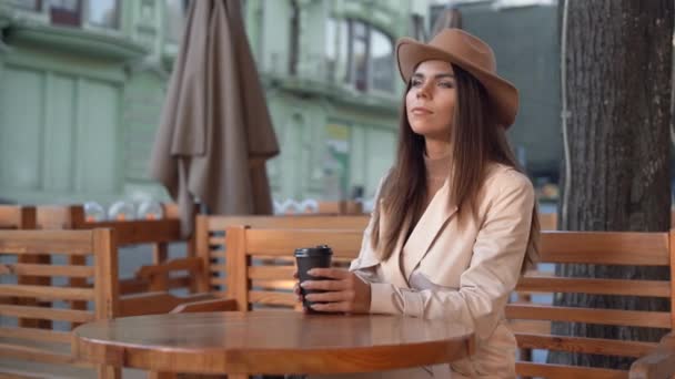 Joven hermosa mujer en un sombrero de fieltro se sienta en una mesa en un café en la calle. beber café, taza de papel desechable — Vídeos de Stock