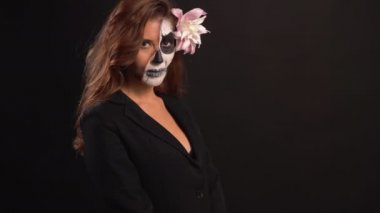 Karanlık bir arka planda Cadılar Bayramı için makyajlı genç bir kadının portresi. Ayakta durur ve sakince bakar. Metni kopyala