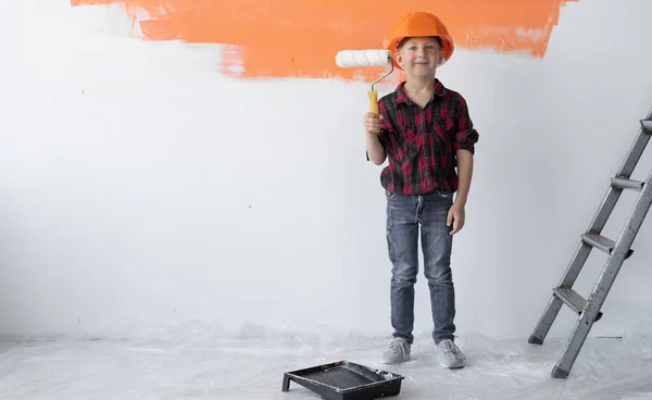 Un niño pequeño vestido con un casco de seguridad naranja sostiene un rodillo en sus manos. Concepto de renovación del hogar. — Foto de Stock