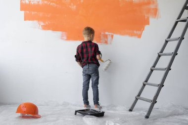 Bir çocuk duvarın yanında duruyor ve onu turuncuya boyuyor. Ebeveynlere yardım etmek.