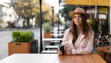 Şapka takmış güzel bir kadın açık hava kafesinde bir masada oturuyor. Tek kullanımlık bir bardaktan kahve iç.