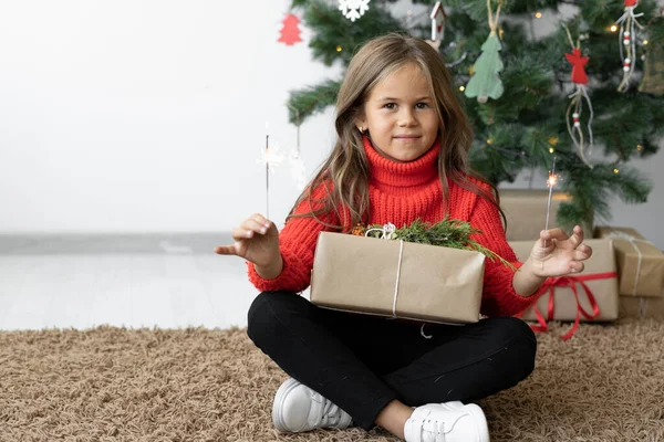 Ein Mädchen europäischen Aussehens sitzt neben dem Weihnachtsbaum. Die Geschenkbox liegt in ihrem Schoß. — Stockfoto