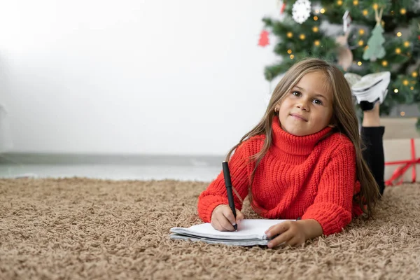 En flicka med europeiskt utseende ligger på golvet nära julgranen. Papper i händerna som skriver ett brev till tomten — Stockfoto
