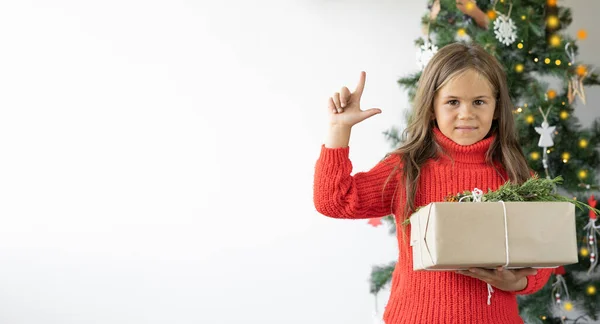 Ein Mädchen steht neben dem Weihnachtsbaum und hält eine Schachtel mit einem Geschenk in der Hand. Zeigefinger zeigt nach oben — Stockfoto