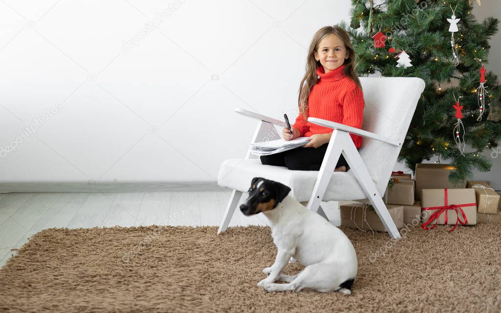smiling girl sits on an armchair near the Christmas tree. The dog jack russell is nearby. Sheet of paper in hands writing a letter to Santa