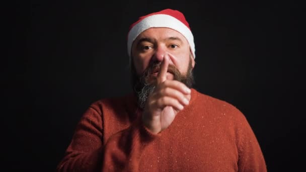 Een goed gevoede man met een baard gekleed in een kerstman hoed steekt een vinger naar zijn lippen en vraagt om stilte. Zwarte achtergrond. Kerstconcept — Stockvideo