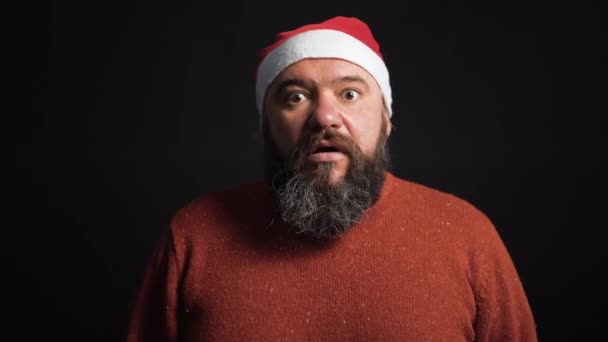 サンタクロースの帽子をかぶった餌を与えられた髭の男は驚き、彼の手のひら黒の背景で顔を覆います。クリスマスのコンセプト — ストック動画