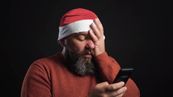 Baculatý vousáč stojí na černém pozadí v klobouku Santa Clause. Když si prohlédne fotku na telefonu, stydí se.. — Stock video