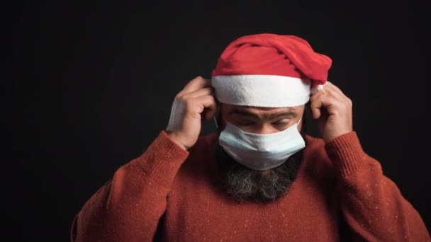Baculatý vousáč stojí na černém pozadí v klobouku Santa Clause. Nosí ochrannou lékařskou masku na obličeji — Stock video