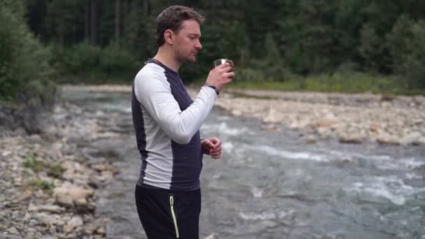 Ein junger Mann steht am Fluss, hält einen Becher Wasser in der Hand und trinkt daraus — Stockvideo