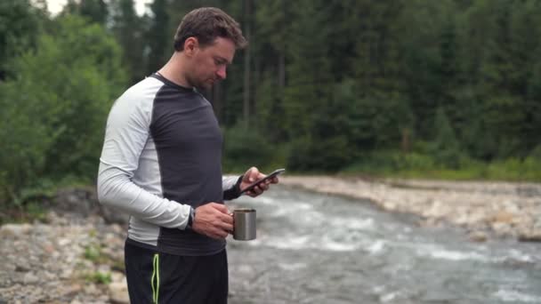Um jovem viajante está perto do rio segurando um copo de água em suas mãos. Digitando uma mensagem em seu smartphone — Vídeo de Stock