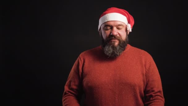 Bearded man on black background wearing santa claus hat unties red ribbon on box — Stock Video