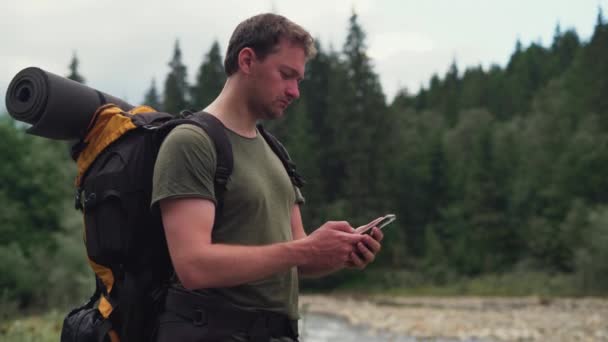 Un turista hombre con una mochila detrás de sus hombros se encuentra cerca del río marca un número en un teléfono inteligente y hace una llamada — Vídeo de stock