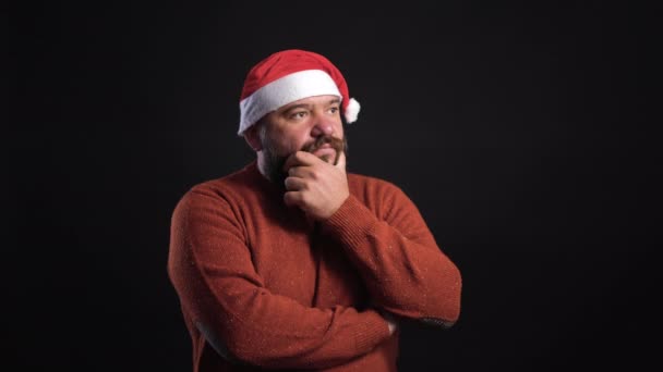 Homem barbudo em um fundo preto vestindo um chapéu de Papai Noel pensa e vem com uma solução para o problema — Vídeo de Stock